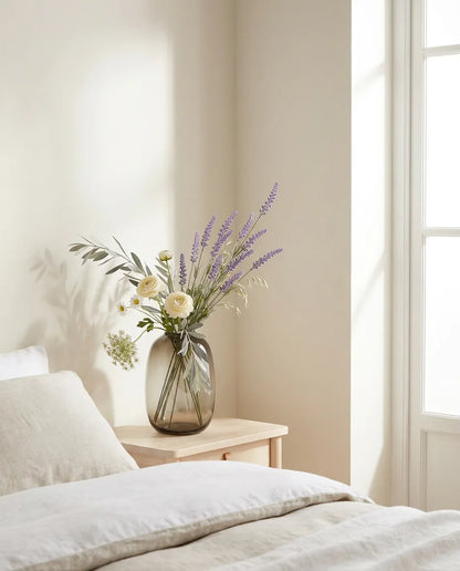 Bouquet artificiel violet et blanc sur une table de chevet en bois clair, avec un linge de lit en lin