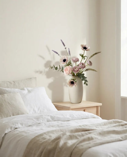 Bouquet artificiel rose et violet sur une table de chevet en bois clair, avec un linge de lit en lin