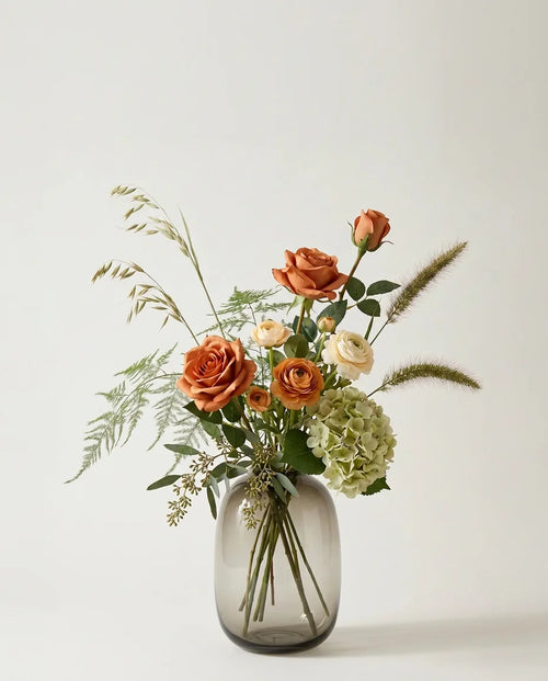 Bouquet artificiel de roses, hortensias et renoncules orange et vert dans un vase en verre ambré