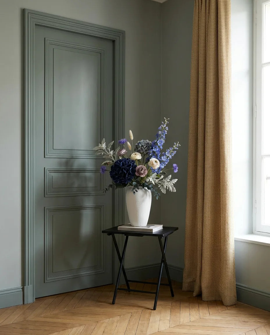 Bouquet artificiel bleu et violet dans une entrée à la porte vert sauge, sur une table d'appoint noire