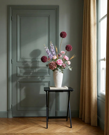Bouquet artificiel rose et violet dans une entrée à la porte vert sauge, sur une table d'appoint noire