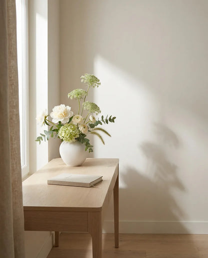 Bouquet artificiel blanc et vert sur un bureau en chêne clair, près d'un rideau en lin