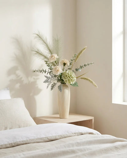 Bouquet artificiel vert et blanc sur une table de chevet en bois clair, près d'un linge en lin