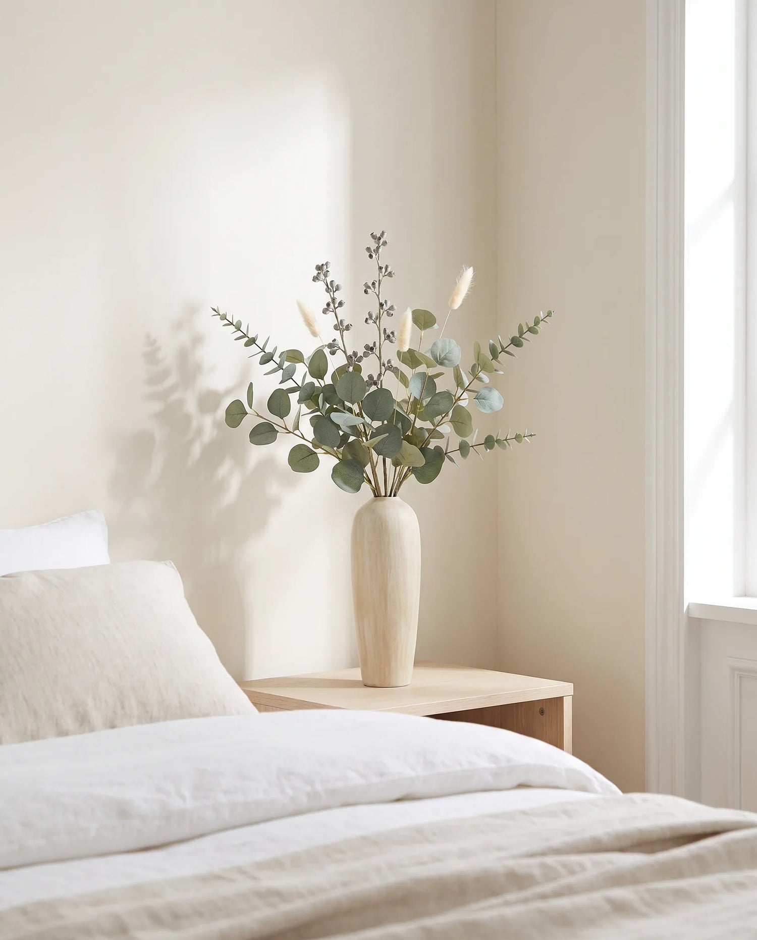 Feuillage artificiel vert-gris sur une table de chevet en bois clair dans une chambre lumineuse