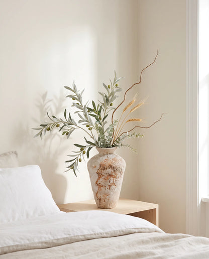 Branchage artificiel vert-olive et doré dans une chambre avec linge de lit en lin et table de chevet en bois clair