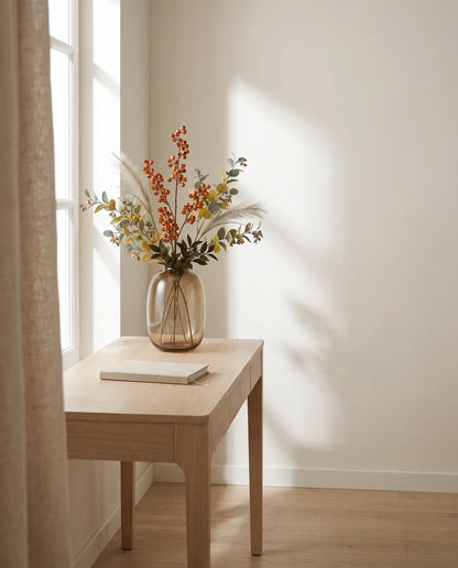 Feuillage artificiel rouille et vert sur une table en chêne clair dans un bureau à la lumière latérale