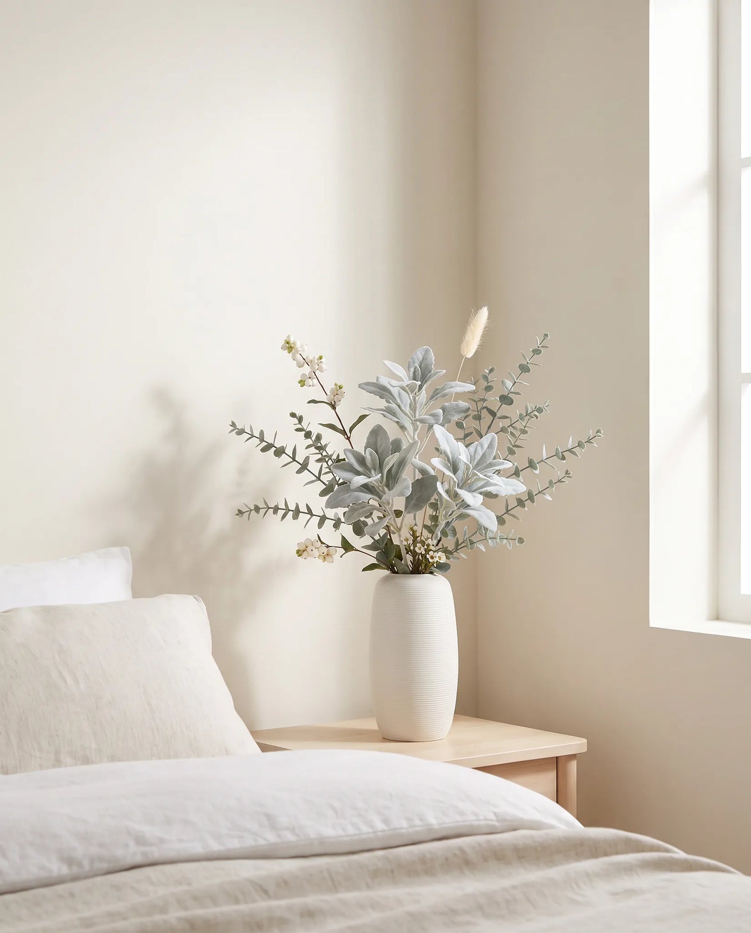 Branches artificielles argent et blanc dans une chambre avec linge de lit en lin et table de chevet en bois clair