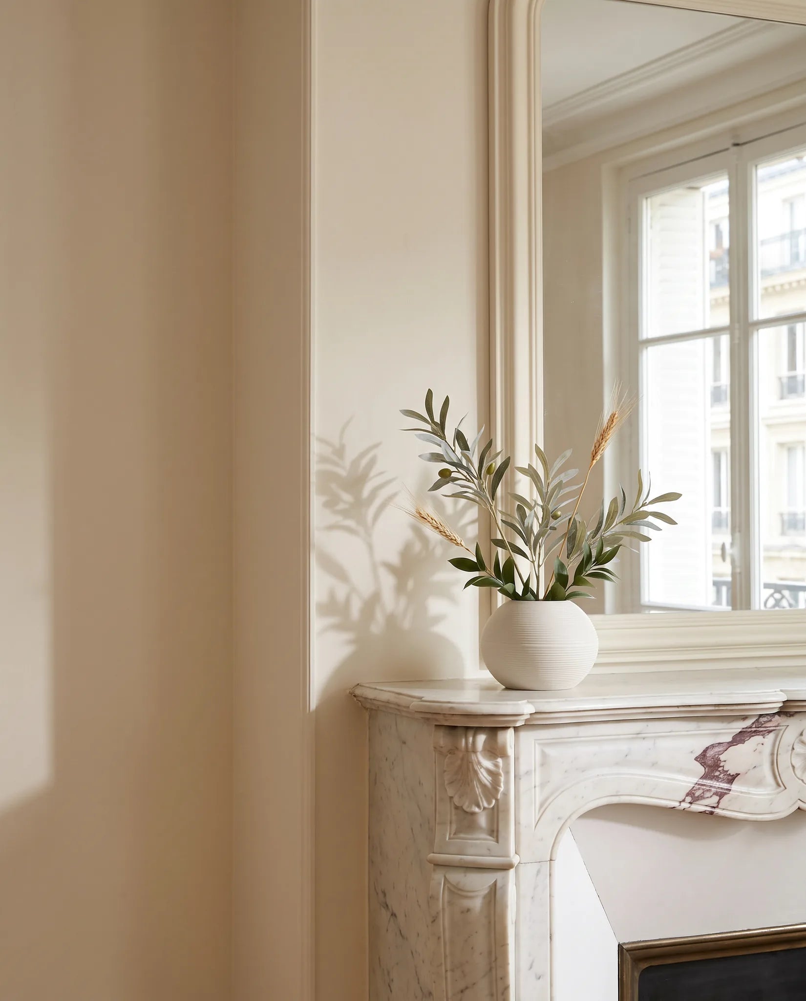 Petite plante artificielle vert-olive et dorée sur une cheminée en marbre près du grand miroir dans un salon parisien rénové